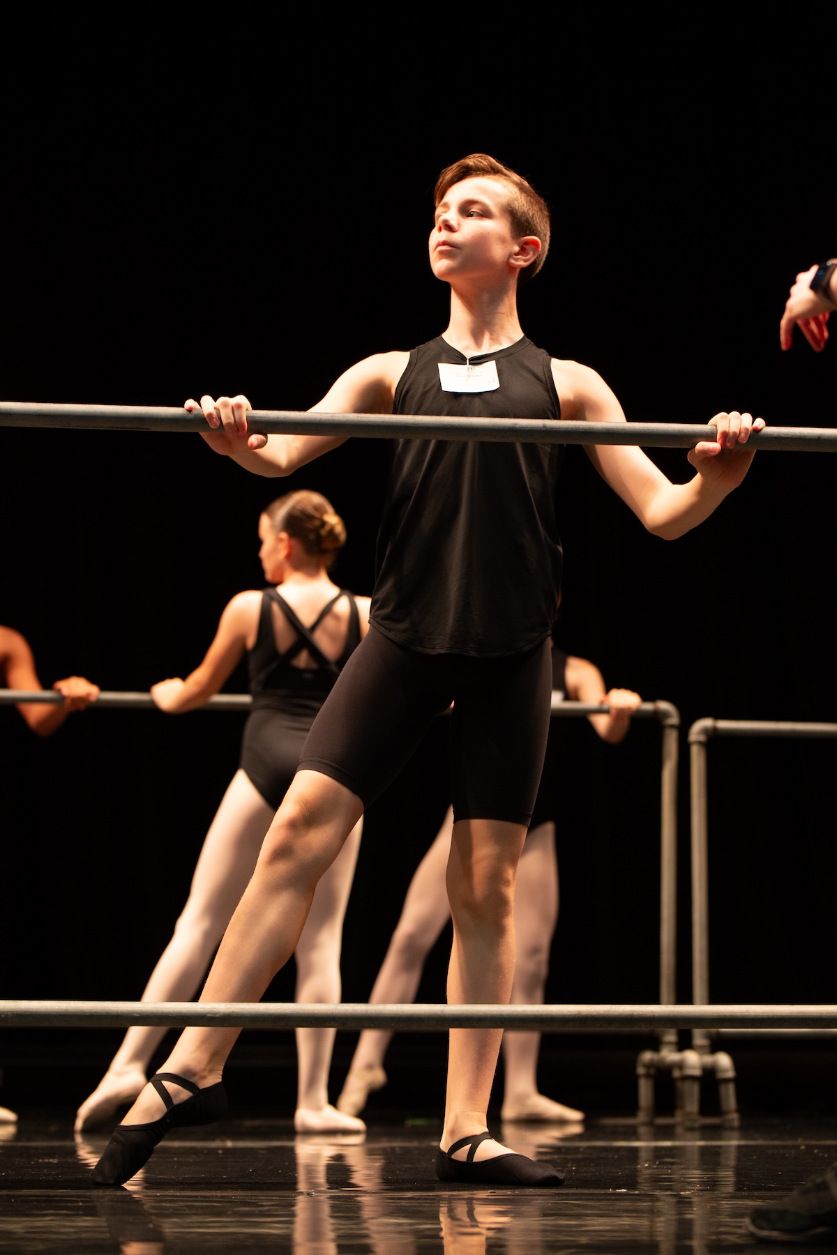 A young male ballet student in a black tank top and shorts performs a barre exercise on a darkly lit stage during a Cecchetti examination, with other dancers visible in the background.