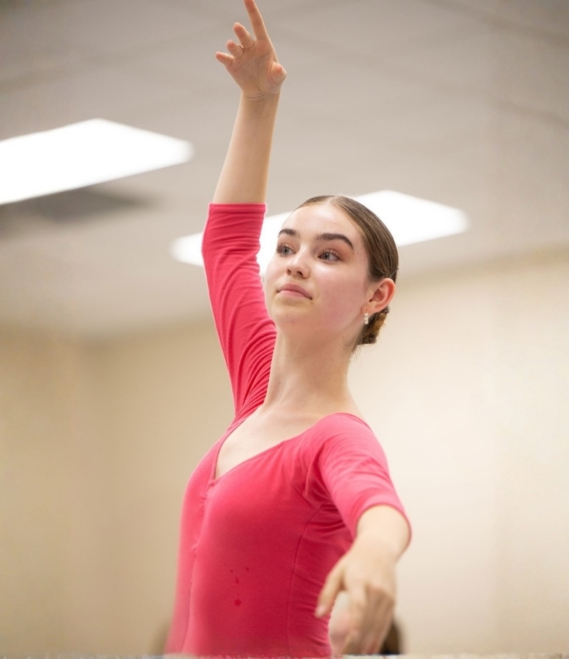 A young female ballet student in a dark pink leotard with hair in a bun raises one arm in a graceful port de bras during a studio class.