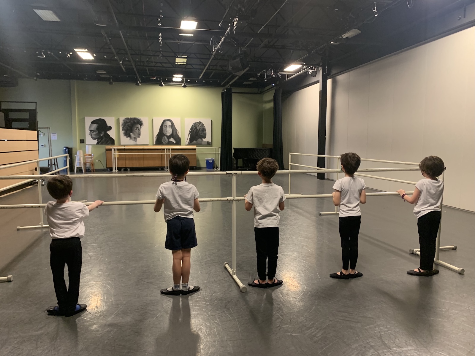 Five young children in white t-shirts stand at a portable ballet barre in a dance studio, seen from behind as they begin an introductory Cecchetti ballet class.